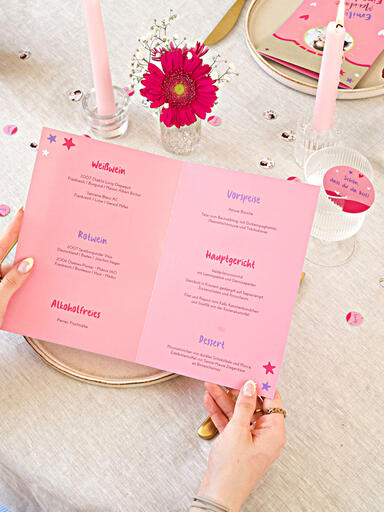 Hände halten eine aufgeklappte Menükarte zur Einschulung gestaltet mit bunten Texten und verschiedenen Schriftarten