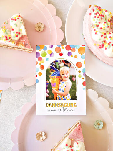 Vorderseite einer Danksagung zur Einschulung mit Foto der Einschulung, bunten Punkten und goldener Veredelung neben Tellern mit bunter Torte